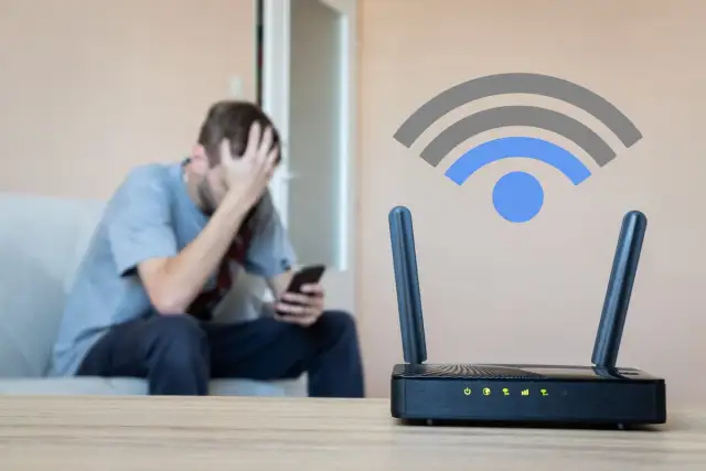 Un homme frustré regarde son téléphone, tandis qu'un routeur wifi est en premier plan. C'est quoi le wifi ? Une connexion sans fil essentielle.