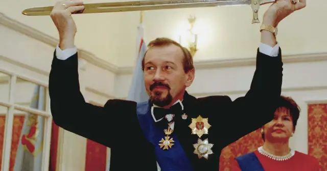 Mężczyzna w czarnym garniturze i orderach trzyma miecz nad głową. Obok kobieta w czerwonej sukni.