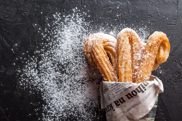 Churros z czekoladą kontra pączki z lukrem? Te złociste churros, posypane cukrem pudrem, kuszą na czarnym tle.