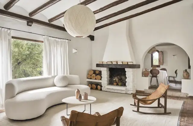 Salón acogedor con sofá curvo blanco, chimenea de piedra y mecedora de cuero. Un ejemplo de casas de campo sencillas y bonitas.