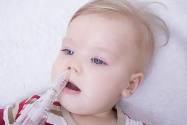 Niemowlę z niebieskimi oczami i blond włosami, któremu odciąga się katar za pomocą aspiratora.