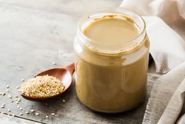 Słoik z gładkim, kremowym tahini obok drewnianej łyżki pełnej nasion sezamu. To właśnie tahini co to jest, czyli pasta z sezamu.
