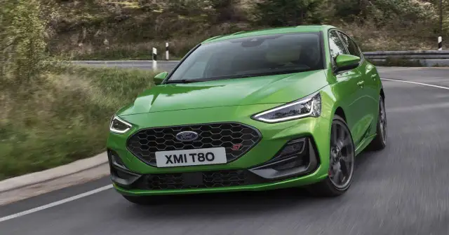Jaskrawozielony Ford Focus 2025 ST z charakterystycznym grillem i światłami LED pokonuje zakręt na malowniczej, leśnej drodze.