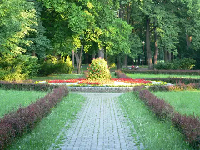 Kamienista ścieżka prowadzi przez park z kwitnącymi rabatami kwiatowymi i zielenią.