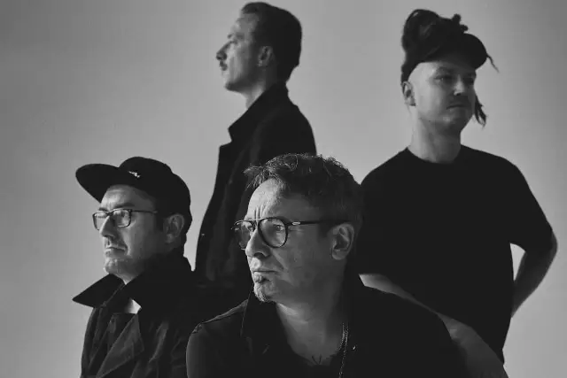 Czterech mężczyzn w czarnych ubraniach, spięty black metalowy zespół, z różnymi fryzurami i okularami, pozuje do zdjęcia.