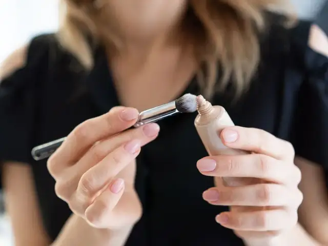 Dłonie z perfekcyjnym manicure nakładają pędzelkiem dr irena eris podkład rozświetlający na pociągłą tubkę.
