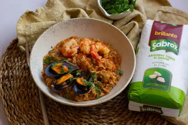 Un plato de arroz con gambas congeladas y mejillones, decorado con perejil fresco. Al lado, un paquete de arroz redondo Sabroz.