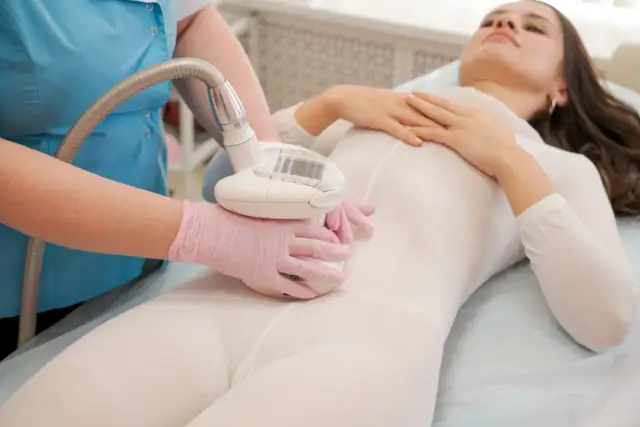 Zabieg endermologii: czy boli? Kobieta w białym kombinezonie leży na łóżku, podczas gdy kosmetyczka wykonuje masaż specjalnym urządzeniem.