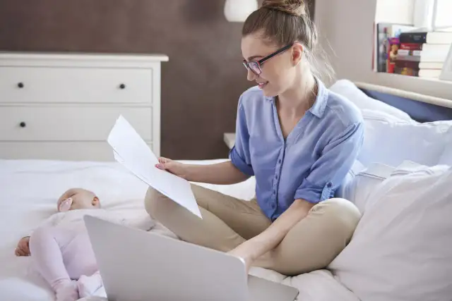 Kobieta na macierzyńskim pracuje przy laptopie, podczas gdy dziecko śpi obok.