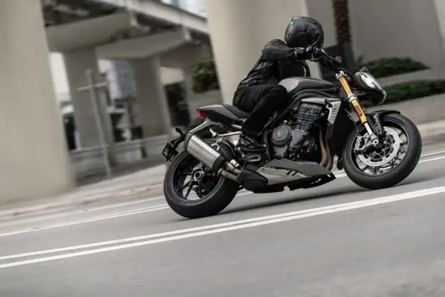Motocyklista w kasku i skórzanej kurtce pochyla się na swoim motocyklu Triumph Speed Triple, pokonując zakręt na miejskiej drodze.