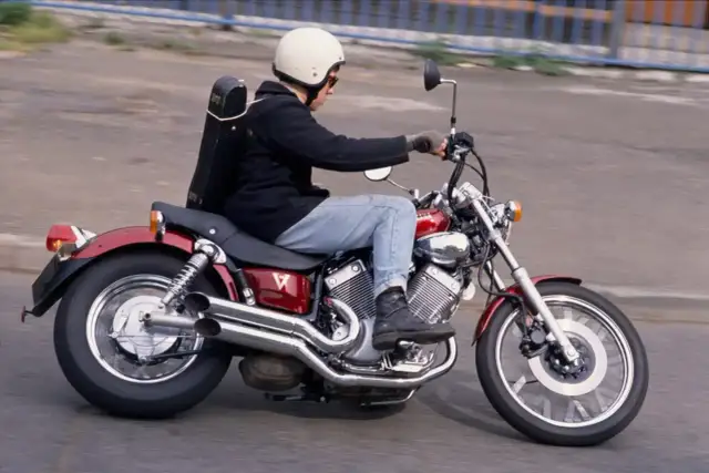 Motocyklista w kasku jedzie na czerwonym motocyklu Yamaha Virago, z futerałem na instrument muzyczny przypiętym z tyłu.