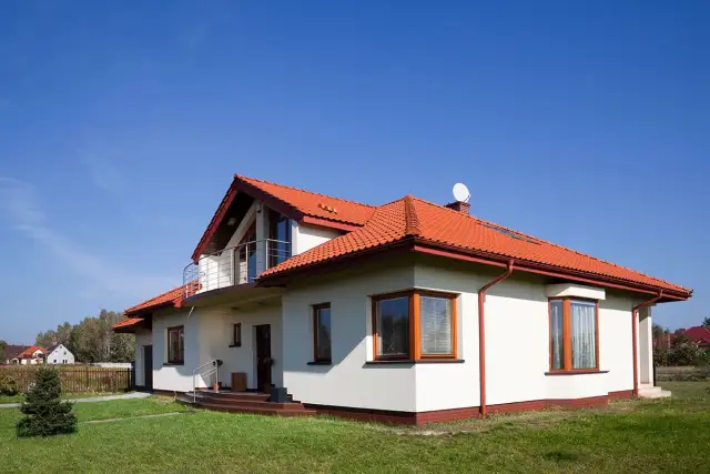 Biały dom z czerwonym dachem i balkonem na tle błękitnego nieba.