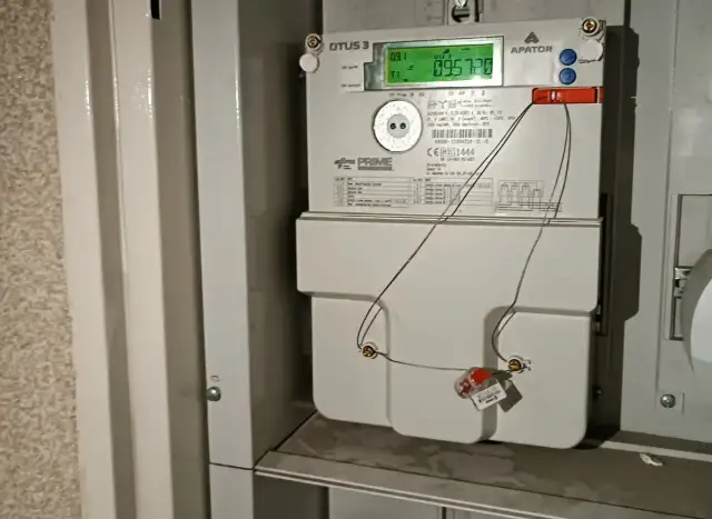 Cyfrowy licznik energii elektrycznej APATOR OTUS 3 z zielonym wyświetlaczem. Widać przewody podłączone do plomby, co może sugerować próby manipulacji, jak oszukać licznik prądu.
