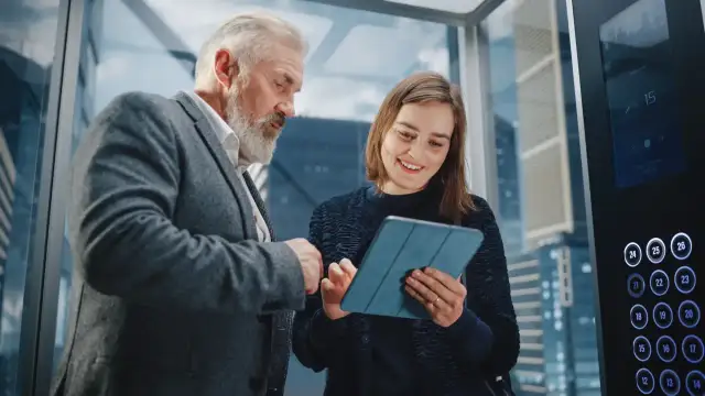 Starszy mężczyzna i młoda kobieta omawiają projekt na tablecie w windzie. To idealny moment na ich elevator pitch.
