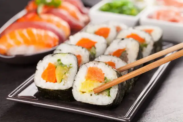 Talerz z sushi i rolkami z łososiem, awokado i marchewką, trzymany pałeczkami.