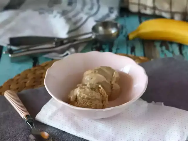 Zwei Kugeln cremiges, veganes Bananeneis in einer rosa Schale, daneben ein Löffel und ein Stück Gebäck. Eine Banane liegt im Hintergrund.