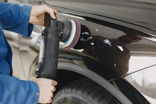 Osoba w niebieskiej kurtce poleruje czarne auto maszyną, pokazując jak usunąć rysy z auta.