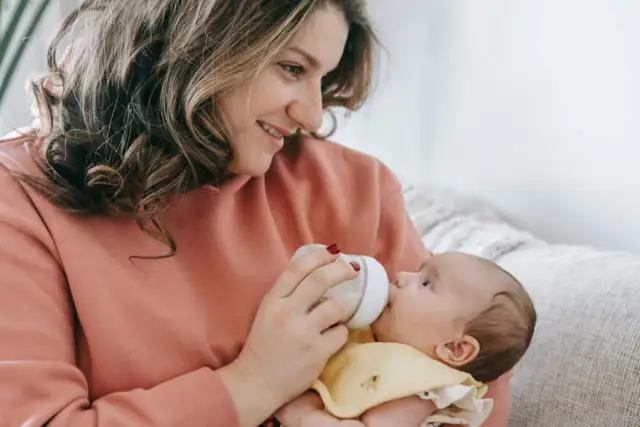 Uśmiechnięta mama karmi butelką dziecko. Warto wiedzieć, ile odciągnięte mleko może stać w lodówce, by zapewnić mu bezpieczeństwo.