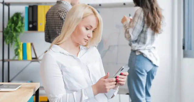 Kobieta z blond włosami korzysta z telefonu, być może używając wyszukiwania głosowego, podczas gdy koledzy pracują przy tablicy.