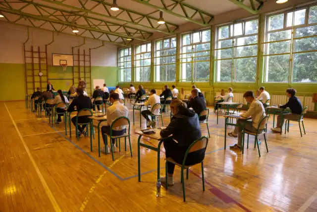 Uczniowie siedzą przy ławkach w sali gimnastycznej, pisząc egzamin.