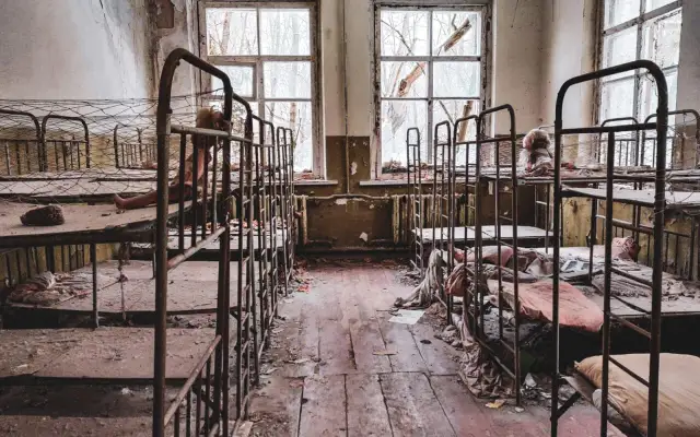 Opuszczony pokój z rzędami zardzewiałych łóżek piętrowych, zniszczonymi oknami i śmieciami na podłodze.