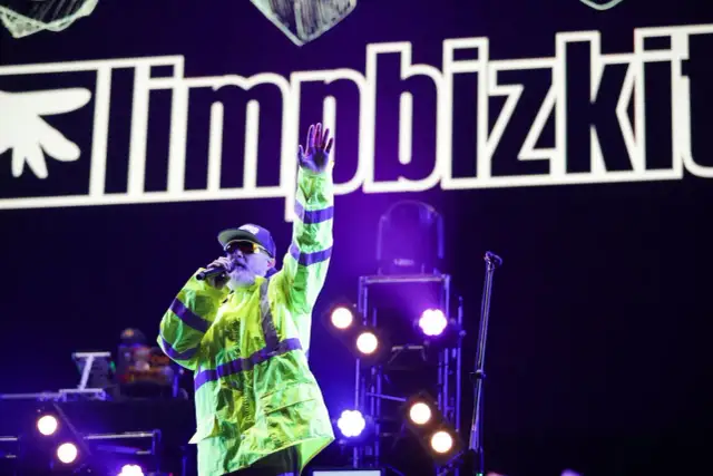 Fred Durst z Limp Bizkit na koncercie, w neonowej kurtce, z ręką w górze. Tło to logo zespołu.