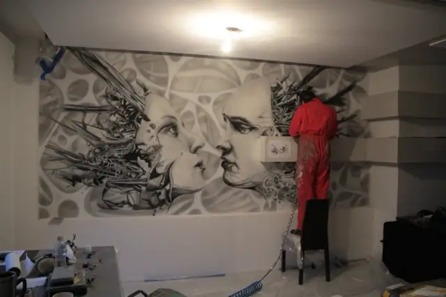 Artysta w czerwonym kombinezonie tworzy malarstwo artystyczne wnętrz – surrealistyczny mural z twarzami i mechanicznymi elementami.
