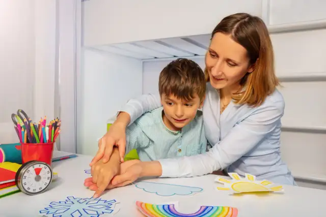 Kobieta pomaga chłopcu w zajęciach plastycznych, tworząc prace na temat pogody. To przykład wsparcia, które może być częścią świadczenia pielęgnacyjnego 2026.
