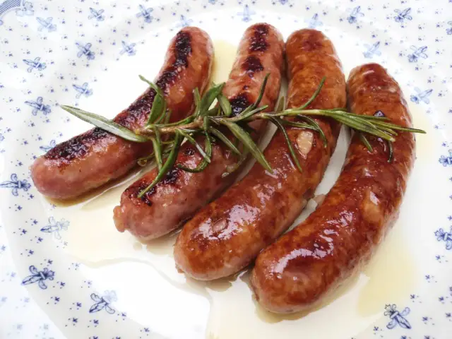 Salchichas doradas en salsa, con romero fresco, servidas en un plato blanco con diseño azul.