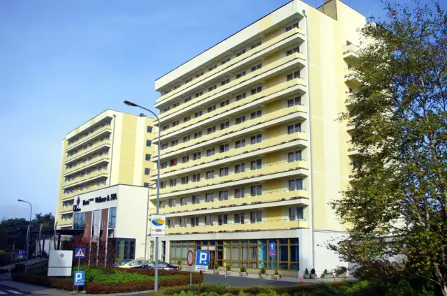 Hotel Vestina w Międzyzdrojach, wysoki, żółty budynek z balkonami, otoczony zielenią.