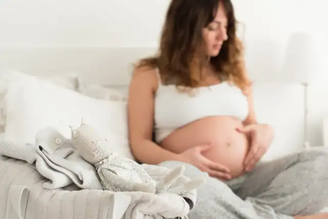Czekając na pierwsze kopnięcia, przyszła mama głaszcze brzuszek. Obok leżą ubranka i przytulanka dla maluszka.