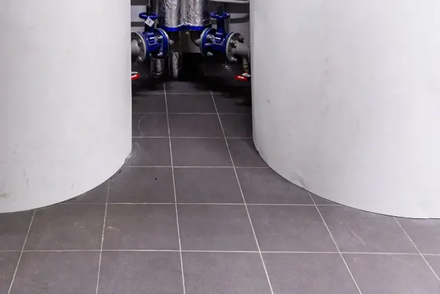 Szare płytki w kotłowni, między białymi zbiornikami. W tle widać instalację hydrauliczną z niebieskimi zaworami.