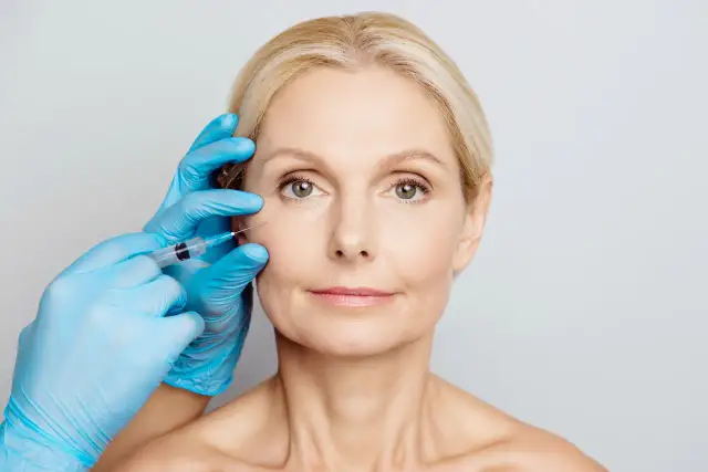 Zabieg z użyciem botoksu, czy botox jest bezpieczny? Dłoń w niebieskiej rękawiczce wykonuje zastrzyk w okolicę oka kobiety.