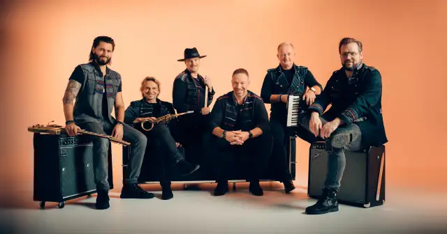Sechs Männer einer Band posieren vor orangefarbenem Hintergrund, einige sitzen, einer steht.