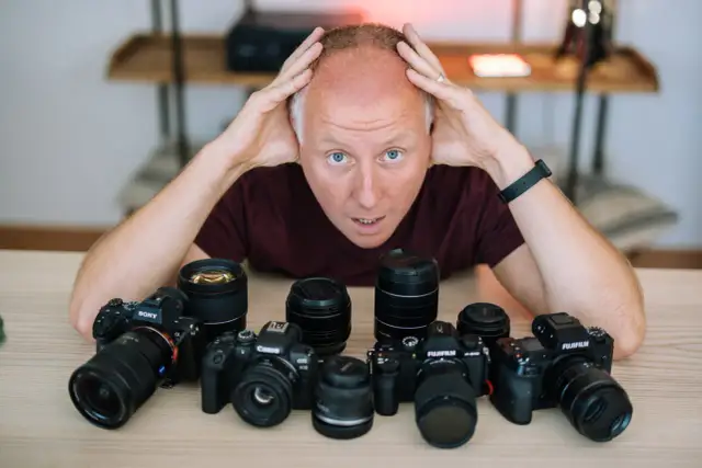 Mężczyzna z rękami na głowie otoczony aparatami różnych marek: Sony, Canon, Fujifilm.