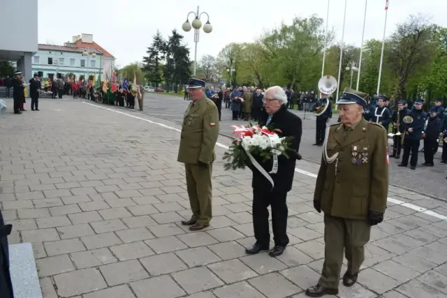 Trzech mężczyzn w mundurach wojskowych i cywilnych, jeden z nich niesie wieniec. W tle parada i orkiestra.