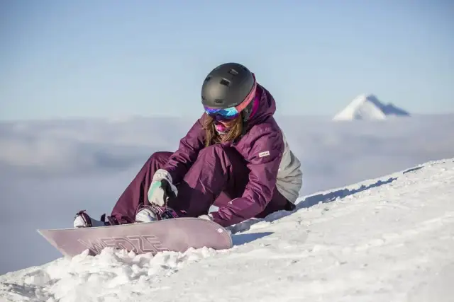 Kobieta w kasku i goglach siedzi na śniegu, zapinając deskę snowboardową.