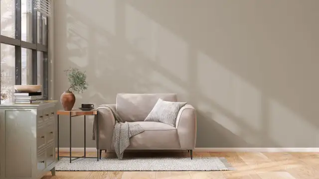 Un sillón beige con manta y cojín, junto a una mesita auxiliar con jarrón y taza. La pared tiene unos colores de pintura neutros, con sombras de luz.