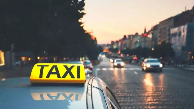 Żółty znak TAXI na dachu samochodu na tle miasta o zachodzie słońca.