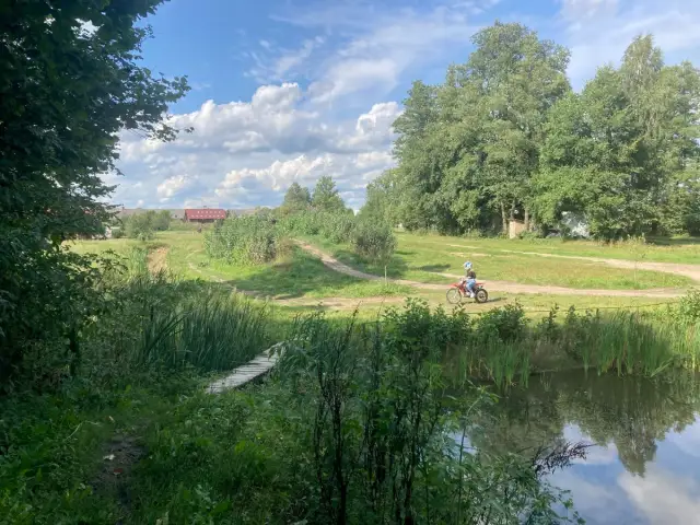 Osoba na motocyklu crossowym jedzie po szutrowej drodze wśród zieleni, obok stawu i drewnianego mostku.