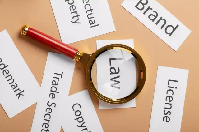 Lupa powiększa słowo "Law" wśród kart z napisami: "Brand", "License", "Copyright", "Trade Secret", "Trademark", "Intellectual Property". To symboliczne przedstawienie analizy prawnej wartości niematerialnych i prawnych.