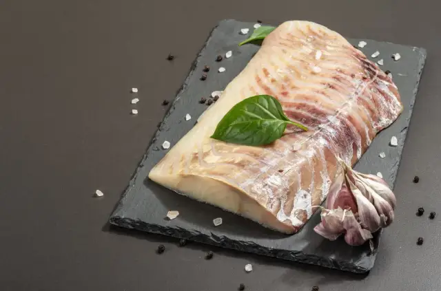 Surowy filet z dorsza czarnego, doprawiony solą i pieprzem, z czosnkiem i listkami bazylii na czarnej desce.
