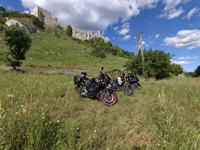 Trzy motocykle stojące na łące z widokiem na ruiny zamku na wzgórzu.