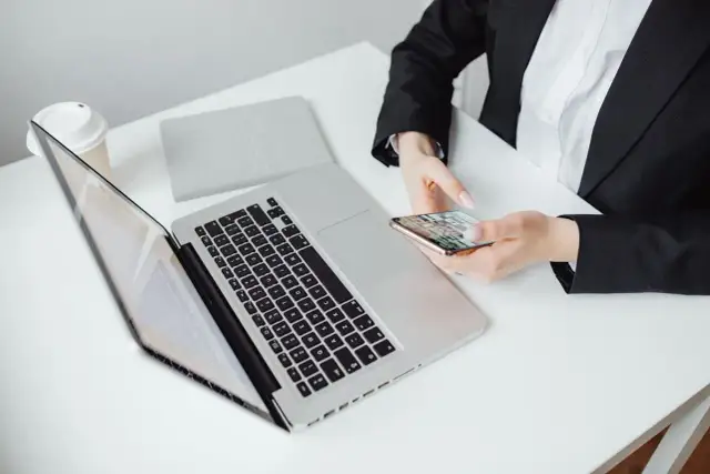 Kobieta w czarnej marynarce trzyma telefon, obok laptop i kubek. Zastanawia się, jak zaadresować pismo do adwokata.