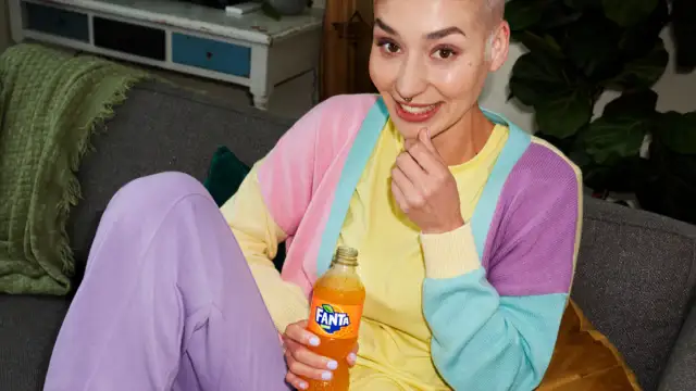 Kobieta w kolorowym swetrze trzyma butelkę Fanta Shokata, siedząc na kanapie.