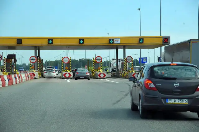 Placówka poboru opłat drogowych z żółtym zadaszeniem, szlabanami i samochodami.