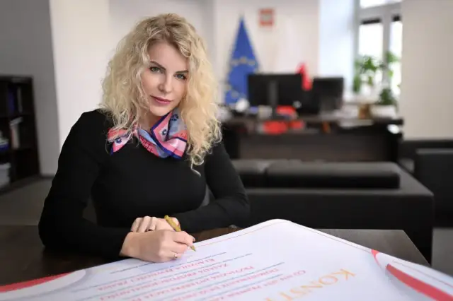 Kobieta z kręconymi blond włosami, w czarnej bluzce i kolorowym szalu, podpisuje dokument. Jest rzecznikiem małych i średnich przedsiębiorców.