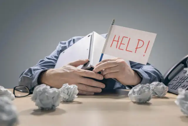Zestresowany mężczyzna pod laptopem z flagą "HELP!" zastanawia się, ile wzrosną raty kredytów. Wokół niego papierowe kule.