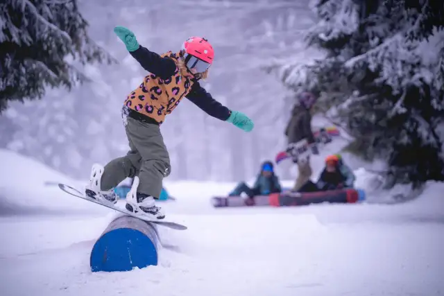 Dziecko na snowboardzie balansuje na rurze w spindleruv mlyn snowpark. W tle inni snowboardziści.