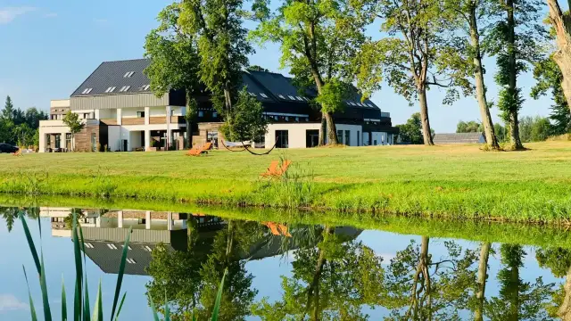 Nowoczesny budynek hotelowy nad jeziorem, z leżakami na trawie i odbiciem w wodzie.